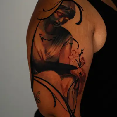 Tattoo on the outer upper arm of a statue of a woman in grey stone tones with red eyes, looking down and cradling a small bunch of red-outlined flowers, framed by bold sweeping black brushstrokes, on melanated skin.