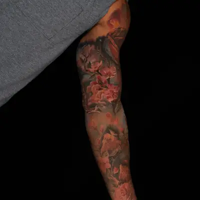 Color realism full sleeve on melanated skin: dense pink cherry blossoms and branches on the forearm with two small birds perched among them, petals drifting upward into a dark cosmic background on the upper arm.