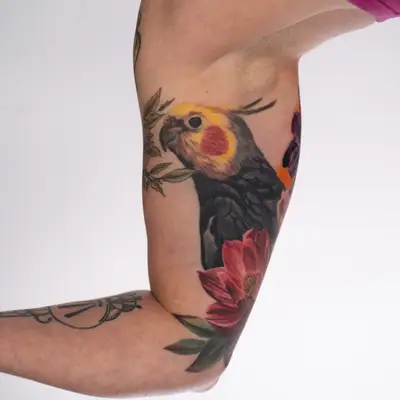 Healed color realism tattoo on the inner bicep of a grey cockatiel with a yellow face and orange cheek patch, surrounded by a deep red flower, green leaves and purple and orange flowers wrapping toward the back of the arm.