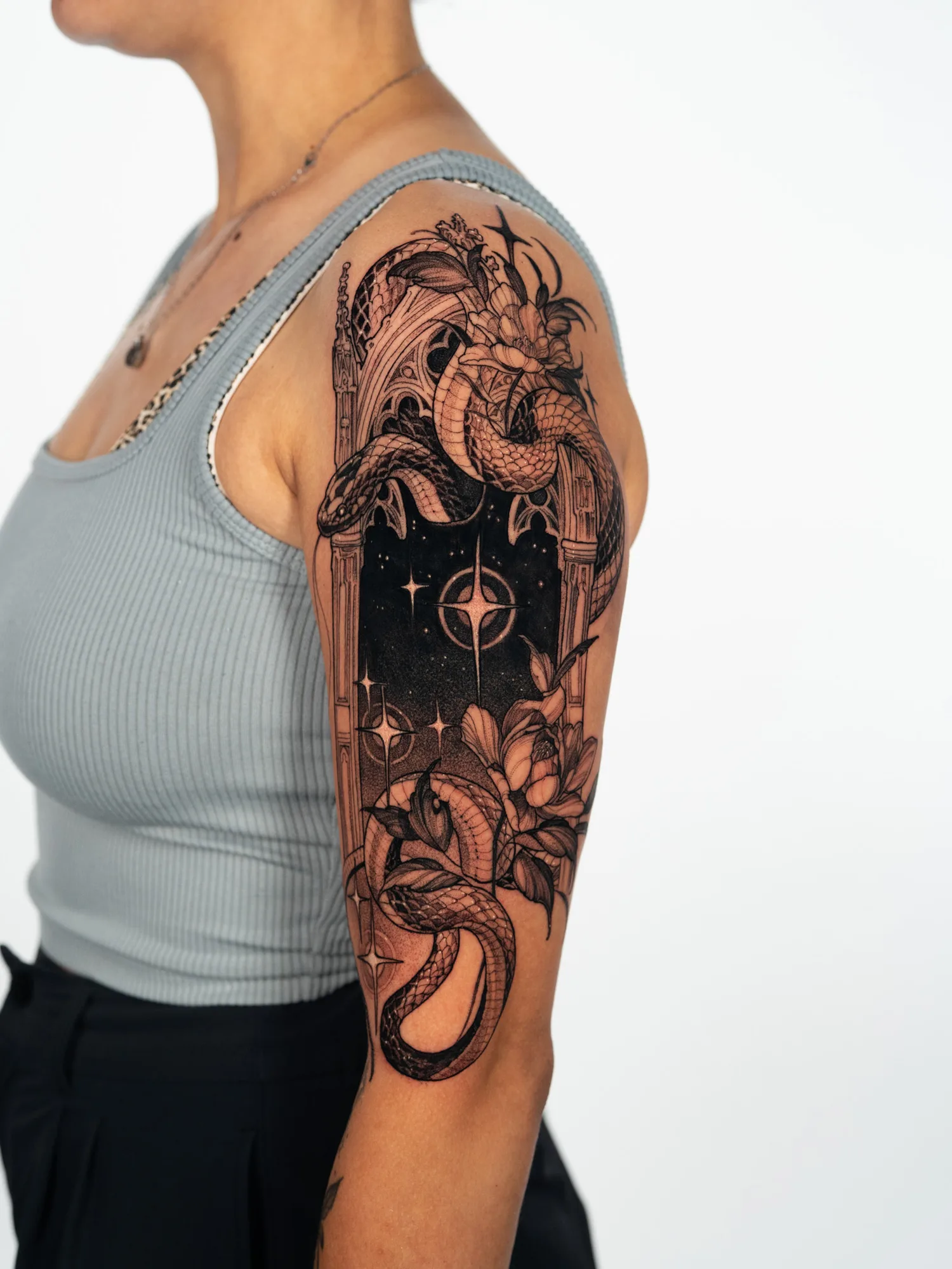 Black and grey tattoo on the upper outer arm of a snake winding through a gothic cathedral window that frames a starry night sky, with full-bloom flowers at the top and lower right.