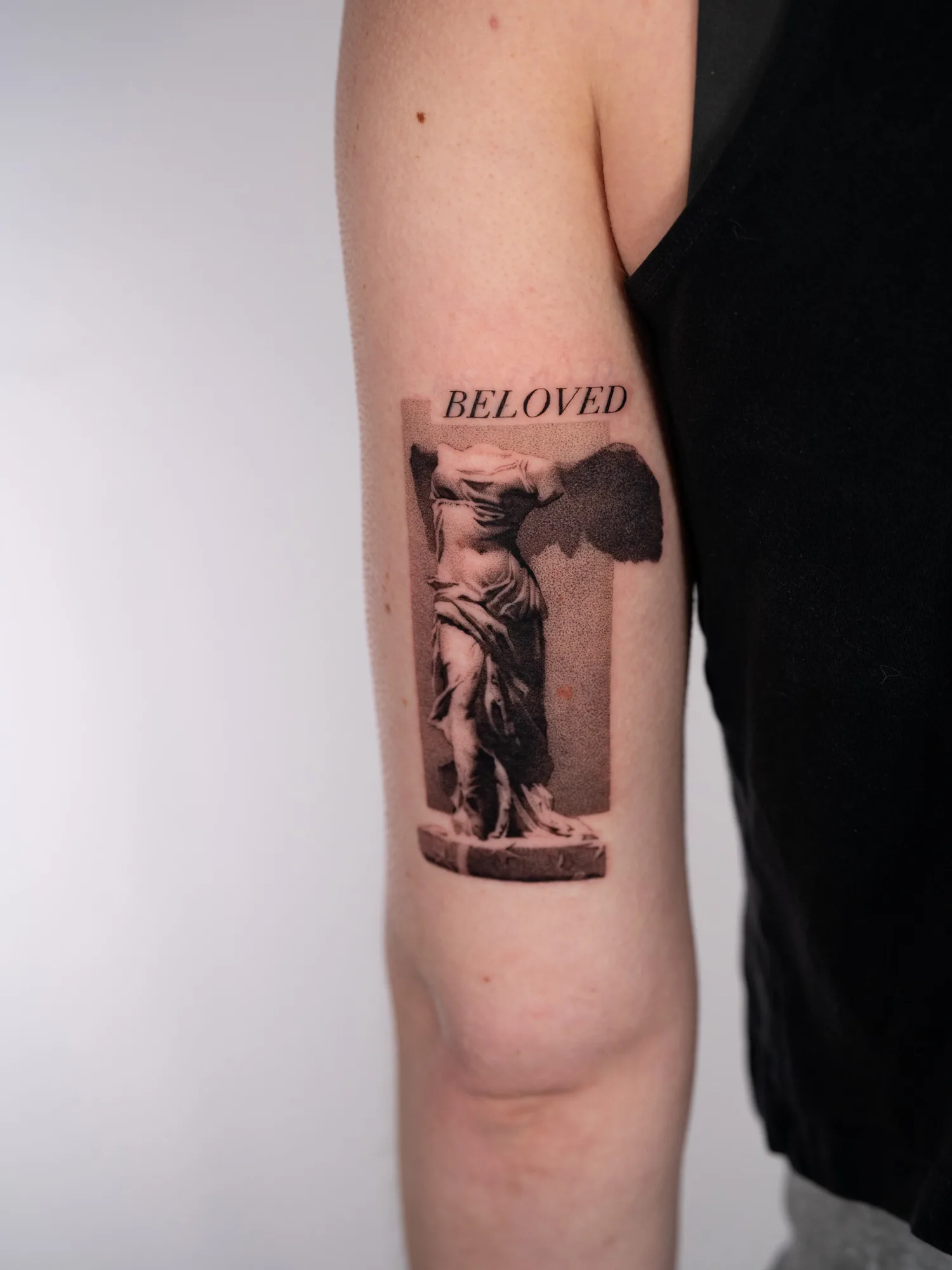 Black and grey realism tattoo on the back of an upper arm of the headless Winged Victory of Samothrace statue on its plinth, set against a stippled grey rectangle with the word BELOVED in serif capitals above.