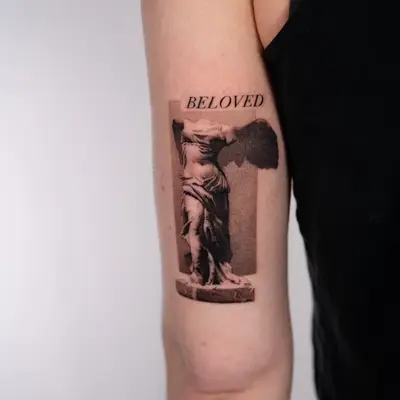 Black and grey realism tattoo on the back of an upper arm of the headless Winged Victory of Samothrace statue on its plinth, set against a stippled grey rectangle with the word BELOVED in serif capitals above.