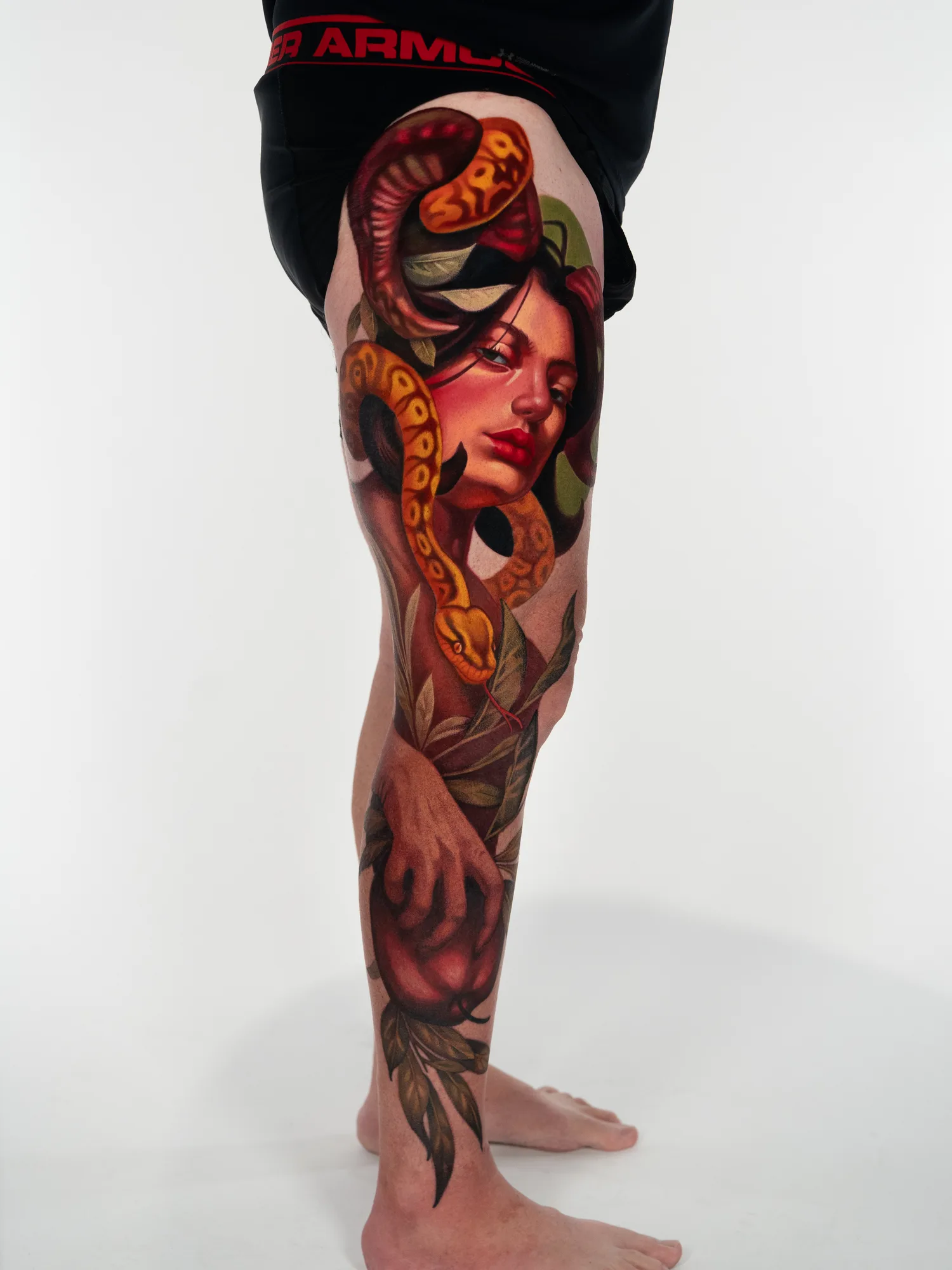 Full-color realism leg sleeve running from the upper thigh to the ankle: a woman's face with red lips surrounded by coiling golden and dark red snakes and green leaves, with a hand gripping a deep red apple on the shin.
