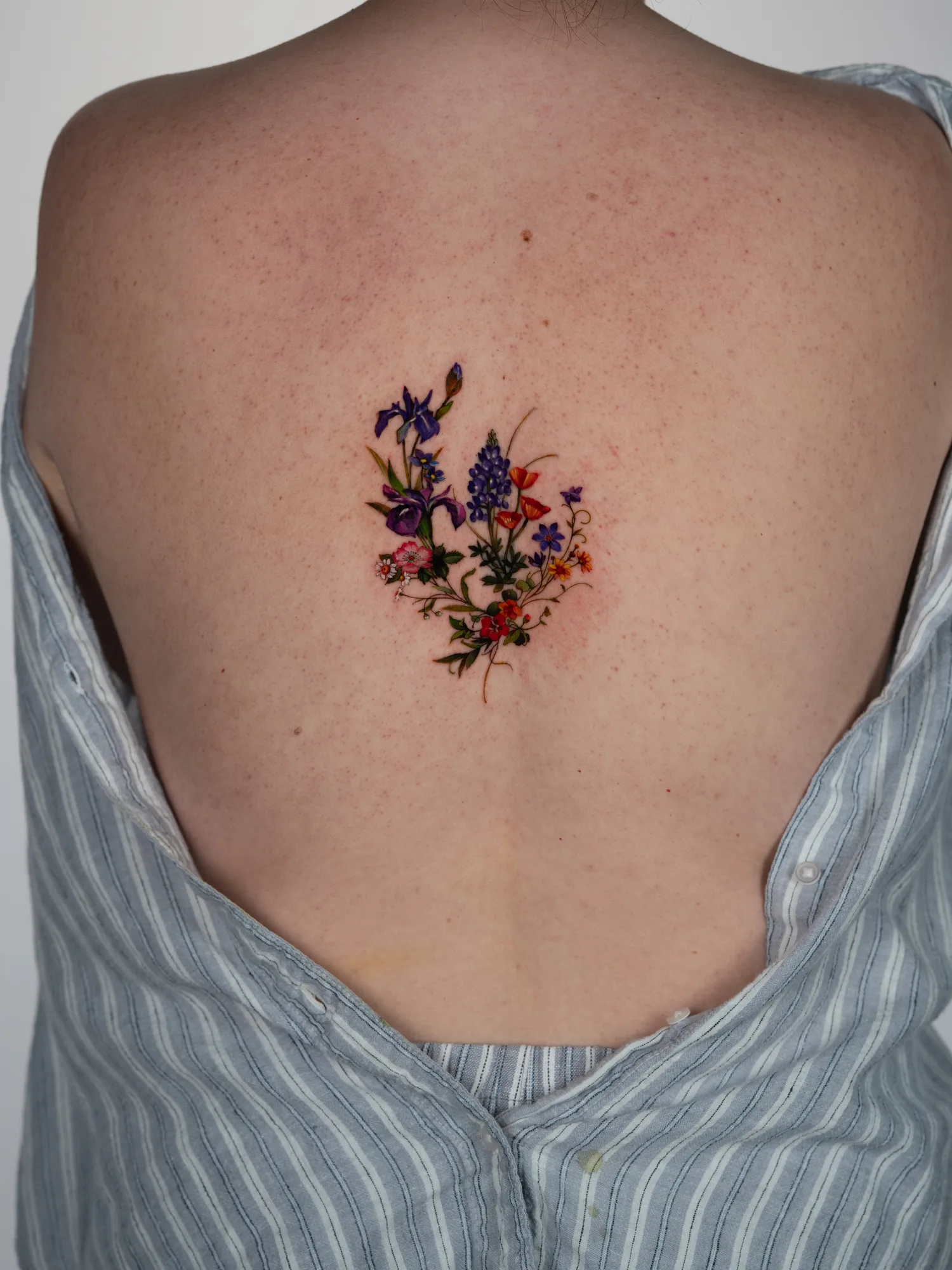 Small color tattoo at the center of the upper back between the shoulder blades: a loose wildflower bouquet of purple irises, blue lupine, orange poppies, small violets and red and pink blossoms with trailing stems.