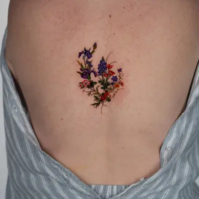 Small color tattoo at the center of the upper back between the shoulder blades: a loose wildflower bouquet of purple irises, blue lupine, orange poppies, small violets and red and pink blossoms with trailing stems.