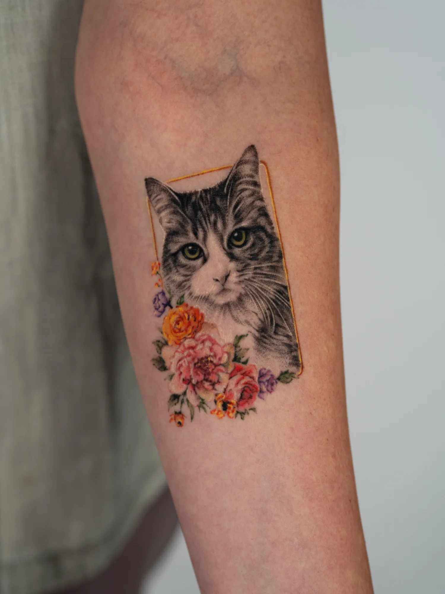 Healed tattoo on the inner forearm: a black and grey realistic portrait of a tabby cat with green-hazel eyes inside a thin gold rectangular frame, whiskers extending past the frame, with a cluster of pink peonies, orange ranunculus, lavender roses and small blooms at the lower left.