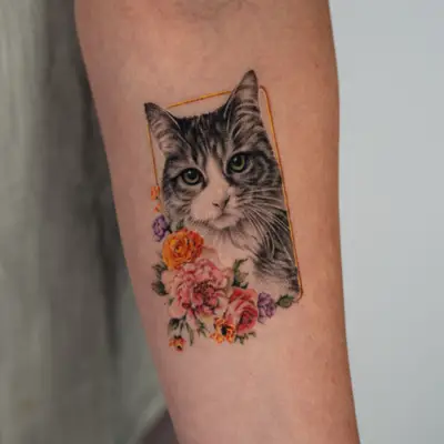 Healed tattoo on the inner forearm: a black and grey realistic portrait of a tabby cat with green-hazel eyes inside a thin gold rectangular frame, whiskers extending past the frame, with a cluster of pink peonies, orange ranunculus, lavender roses and small blooms at the lower left.