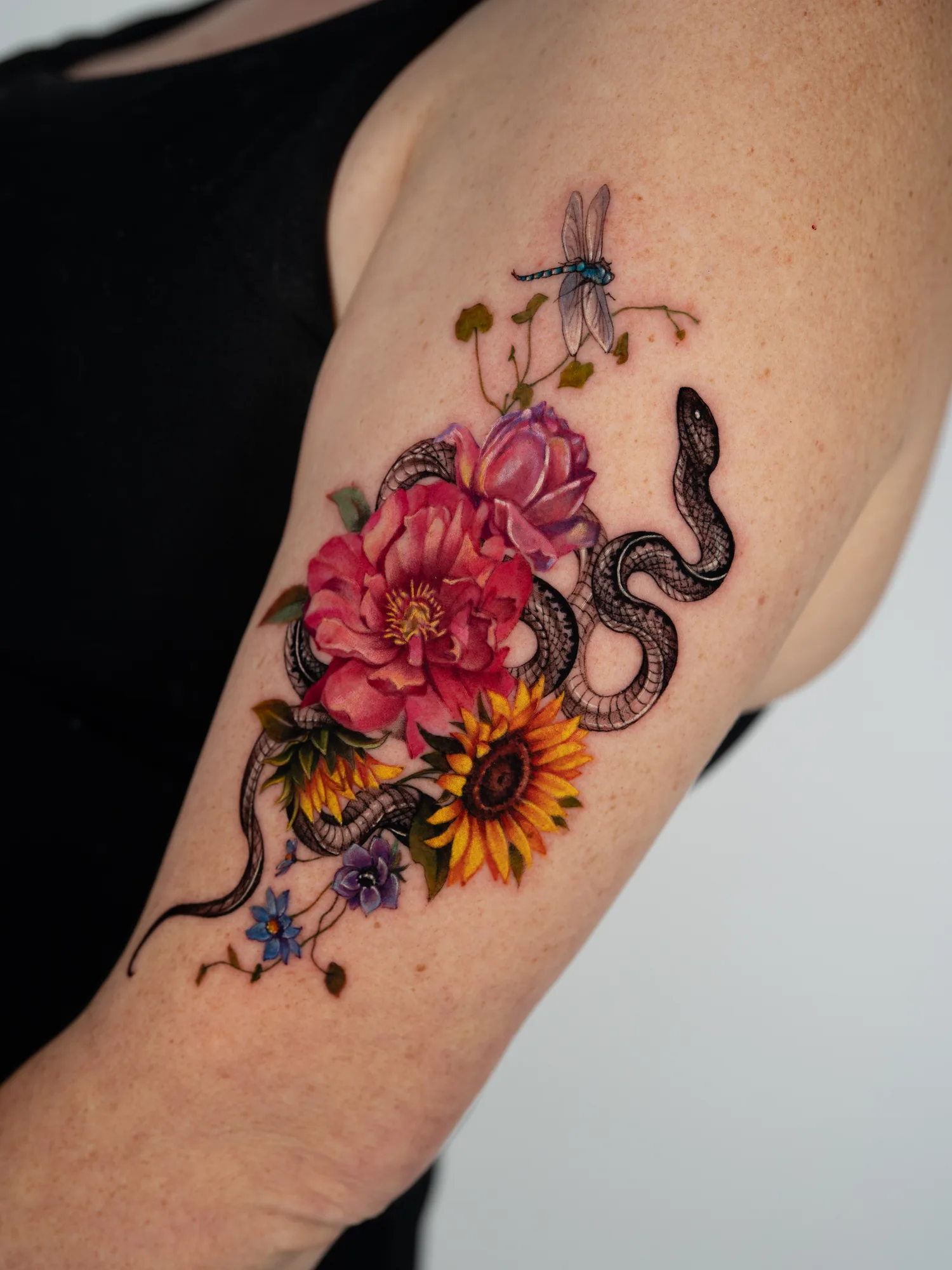 Color realism tattoo on the outer upper arm: a black snake winding through a bouquet of pink and magenta peonies and two sunflowers, with small blue and purple flowers below and a blue dragonfly rising above the arrangement.