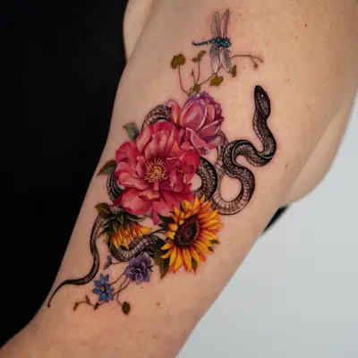 Color realism tattoo on the outer upper arm: a black snake winding through a bouquet of pink and magenta peonies and two sunflowers, with small blue and purple flowers below and a blue dragonfly rising above the arrangement.