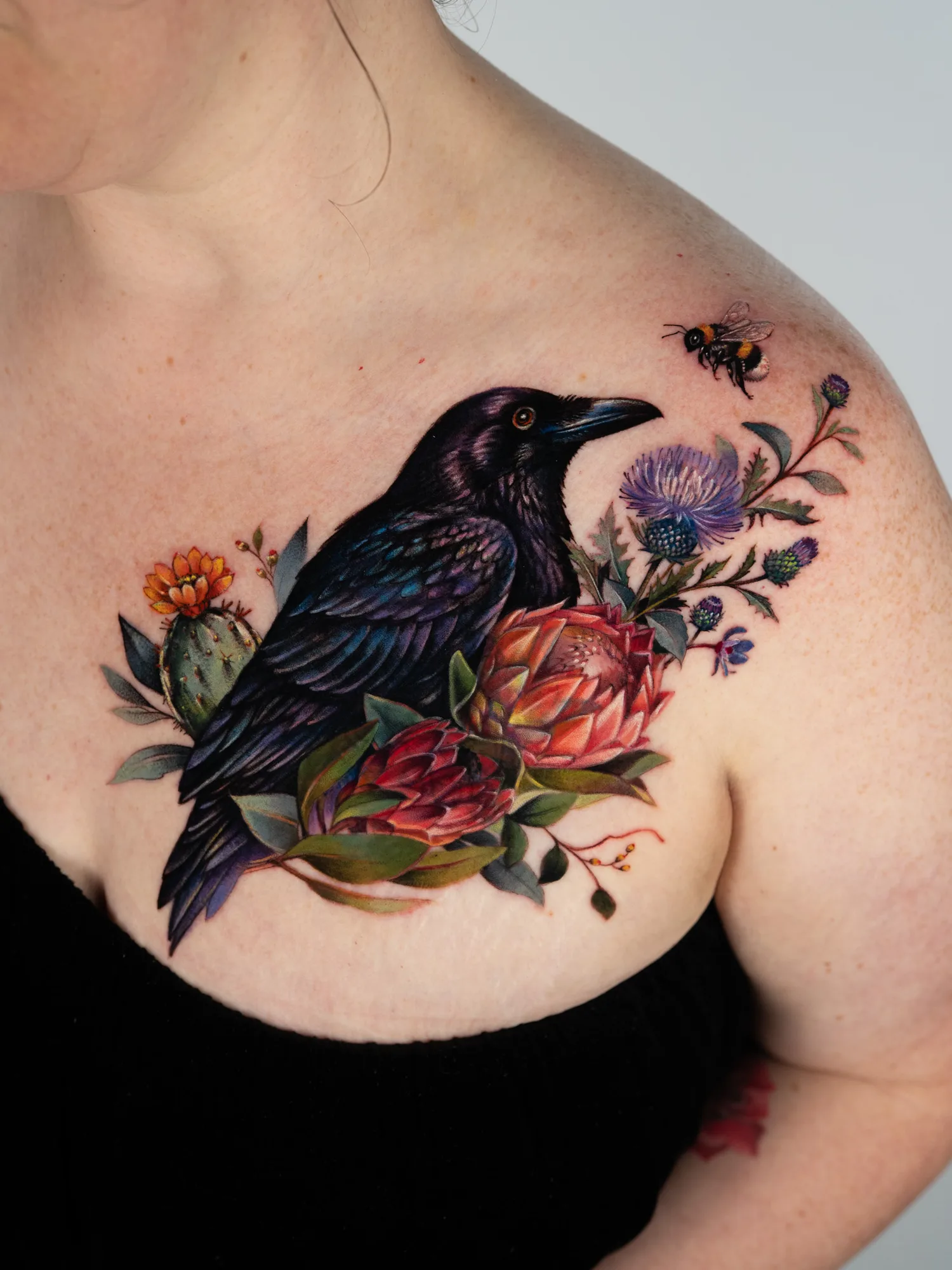 Color realism cover-up across the upper chest and collarbone toward the shoulder: an iridescent black crow with an orange eye among pink and red protea, a flowering prickly pear cactus, purple thistles and eucalyptus leaves, with a bumblebee near the shoulder.