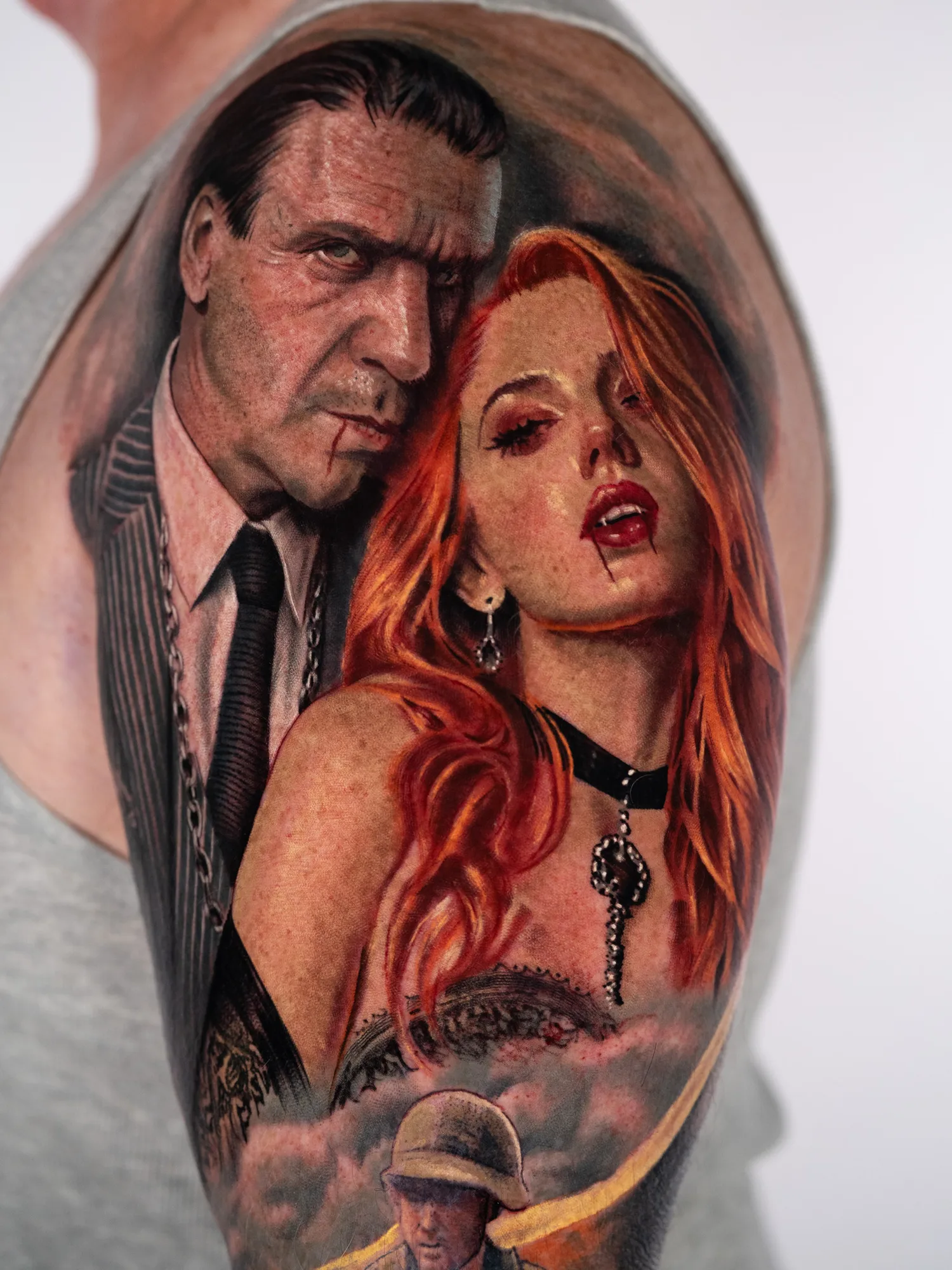 Color realism tattoo on the outer upper arm and shoulder: a stern dark-haired man in a pinstripe suit and chain with blood at his lip, beside a red-haired woman with fangs, blood on her lips, an earring and a black choker, above swirling smoke from which a soldier in a Vietnam-era helmet emerges.