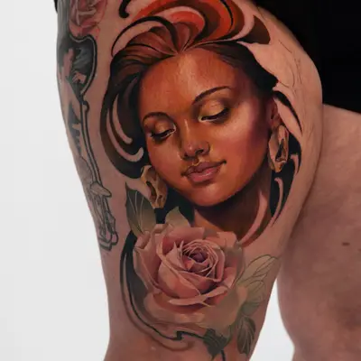 Color tattoo on the thigh of a serene young woman with closed eyes and warm brown skin, framed by sweeping dark and orange hair strokes, with a large soft pink rose beneath her face.