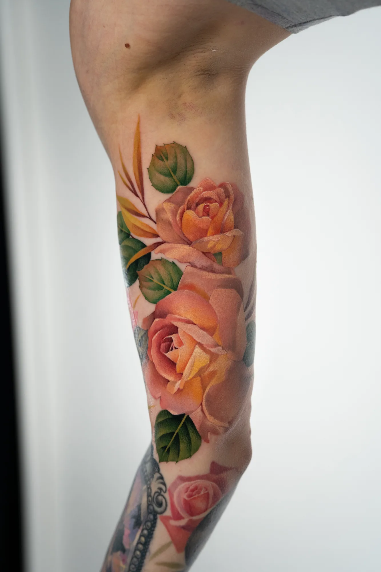 Color tattoo on the inner bicep of two large peach and apricot roses with green leaves and amber fronds, joining an existing sleeve below that includes a pink rose and a jeweled frame.