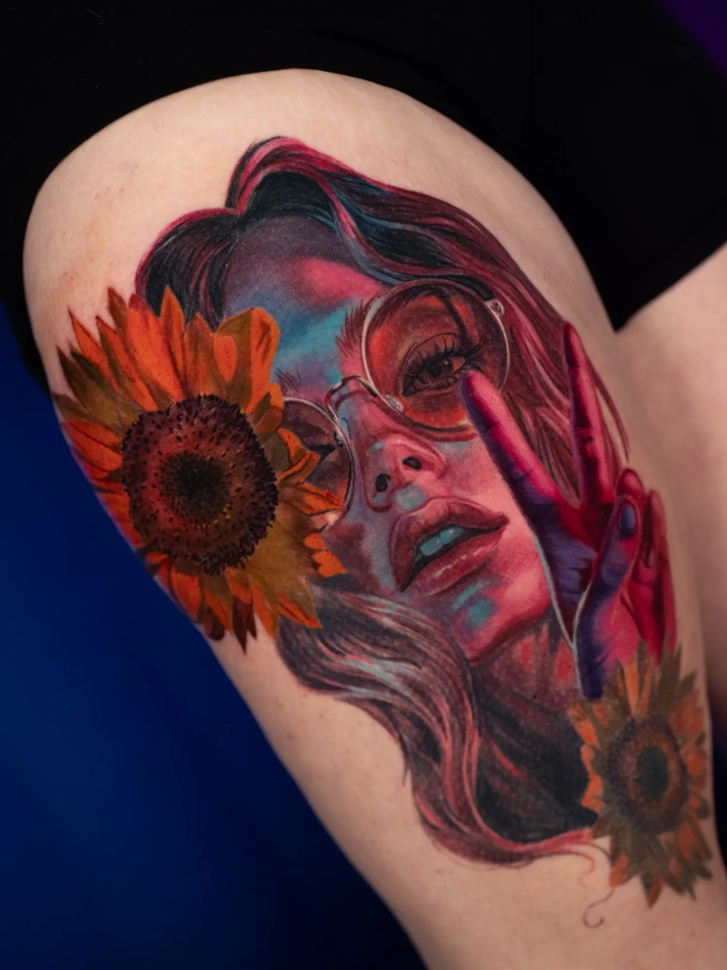 Color tattoo on the front of the thigh of a woman with long wavy hair and round rose-tinted glasses holding a peace sign to her face, her skin washed in pink, purple and teal, with orange sunflowers on either side.
