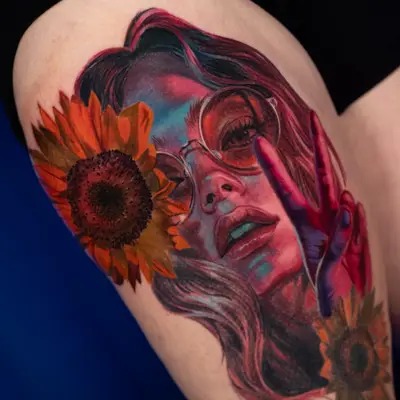Color tattoo on the front of the thigh of a woman with long wavy hair and round rose-tinted glasses holding a peace sign to her face, her skin washed in pink, purple and teal, with orange sunflowers on either side.