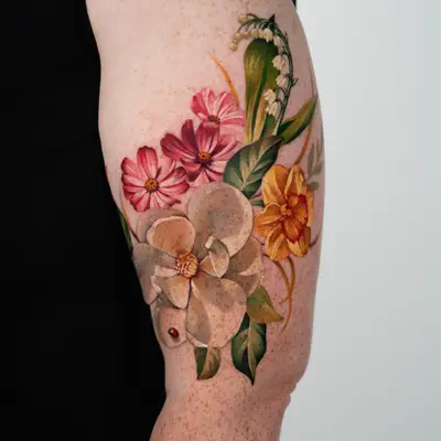 Color tattoo on the outer upper arm of a cream magnolia with a tiny ladybug, pink cosmos, a yellow narcissus and lily of the valley, with green leaves and amber fronds, on freckled skin.