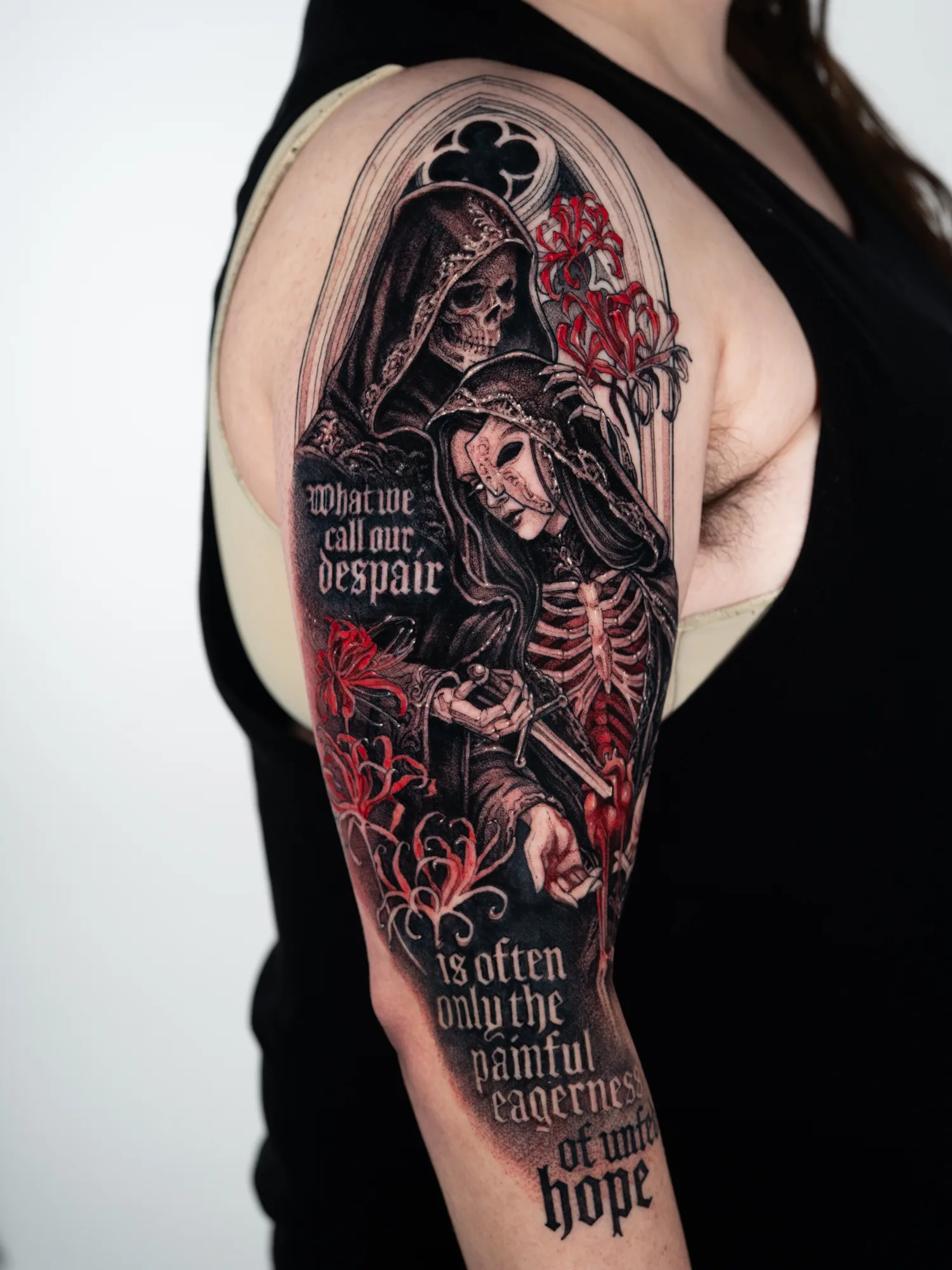 Black and grey tattoo with red accents on the upper arm: a hooded skeletal reaper behind a masked woman holding her own bleeding heart out from an open ribcage, under a gothic arch with red spider lilies, with text reading "What we call our despair is often only the painful eagerness of unfed hope."