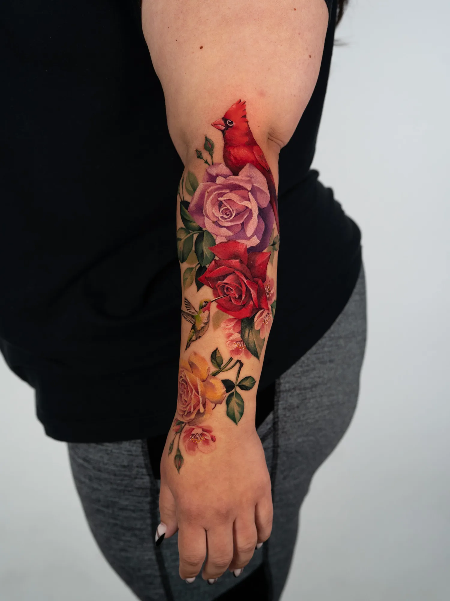 Color tattoo running the full length of the outer forearm: a northern cardinal at the top above lavender and red roses, a ruby-throated hummingbird drinking from a red rose, a yellow rose near the wrist, and pink cherry blossoms throughout.