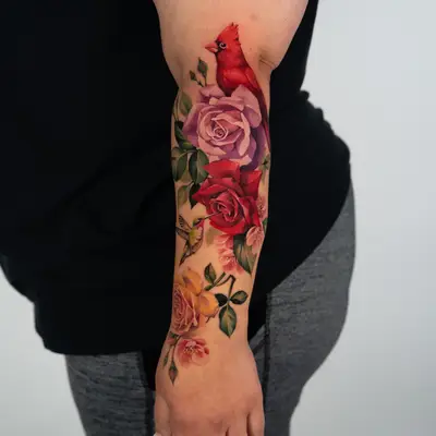 Color tattoo running the full length of the outer forearm: a northern cardinal at the top above lavender and red roses, a ruby-throated hummingbird drinking from a red rose, a yellow rose near the wrist, and pink cherry blossoms throughout.