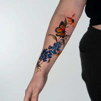 Color tattoo on the forearm, in progress: a cluster of blue Texas bluebonnets rising from the wrist, a monarch butterfly and a black-eyed Susan near the elbow, with green leaves and small sprigs of white flowers.