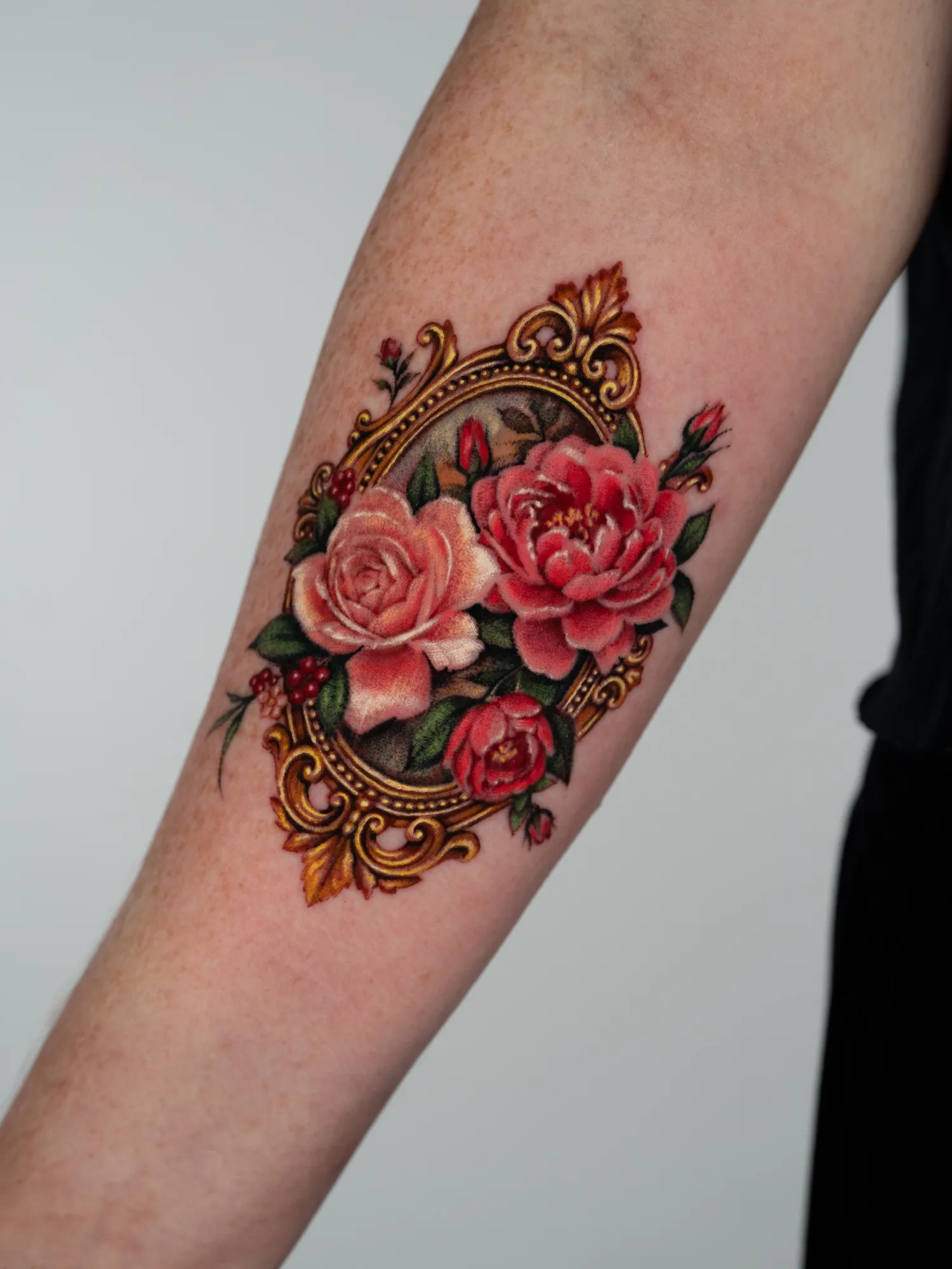 Color micro-realism tattoo on the inner forearm: an oval baroque gold frame with a pale pink rose, a coral peony and a small red rose spilling over it, with rosebuds, red berries and green leaves.