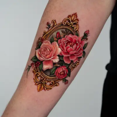 Color micro-realism tattoo on the inner forearm: an oval baroque gold frame with a pale pink rose, a coral peony and a small red rose spilling over it, with rosebuds, red berries and green leaves.