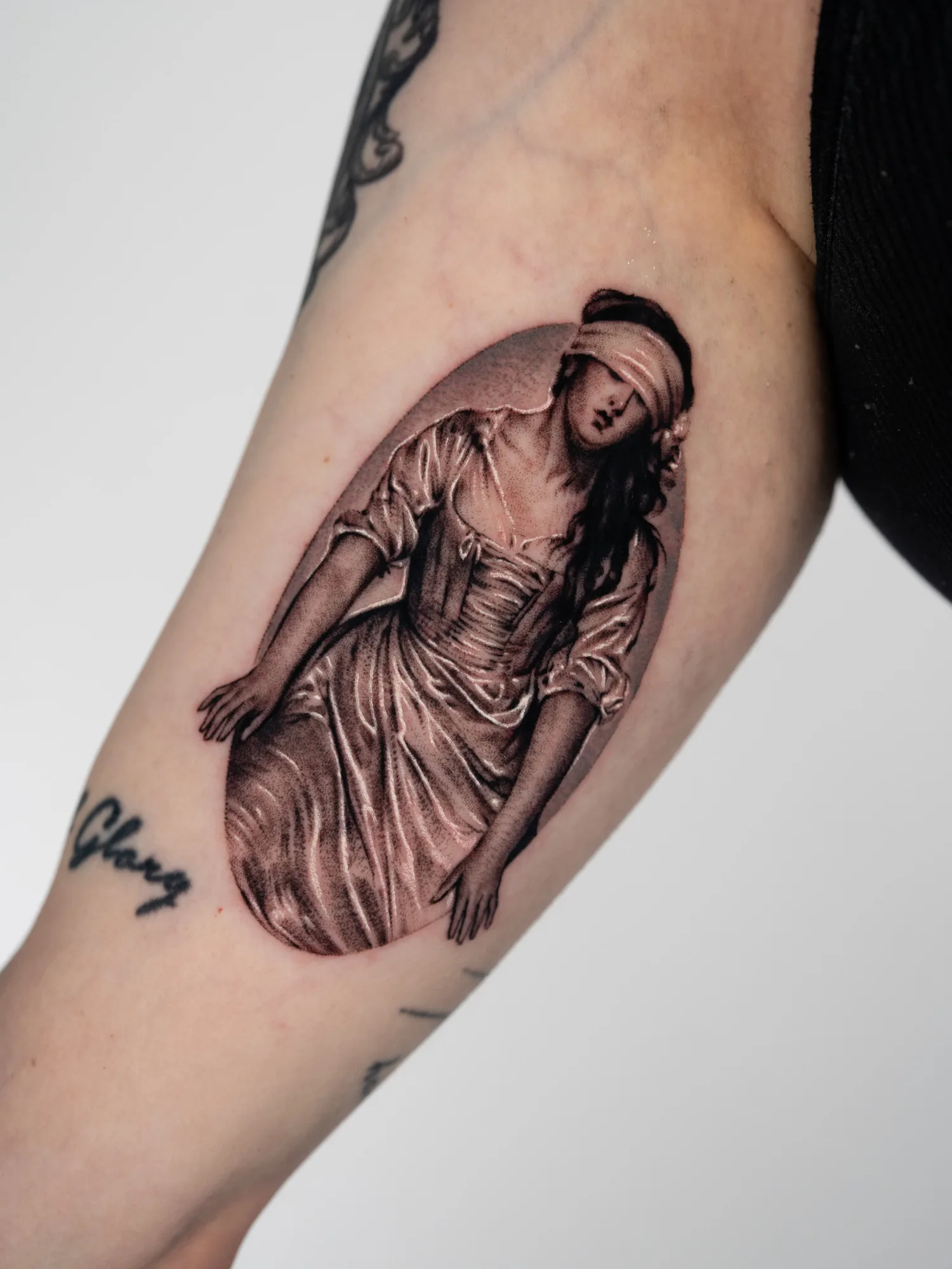Black and grey micro-realism tattoo on the inner upper arm: a blindfolded young woman in a satin gown kneeling and reaching forward with both hands, inside a soft grey oval, after Delaroche's painting of Lady Jane Grey.