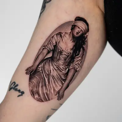Black and grey micro-realism tattoo on the inner upper arm: a blindfolded young woman in a satin gown kneeling and reaching forward with both hands, inside a soft grey oval, after Delaroche's painting of Lady Jane Grey.