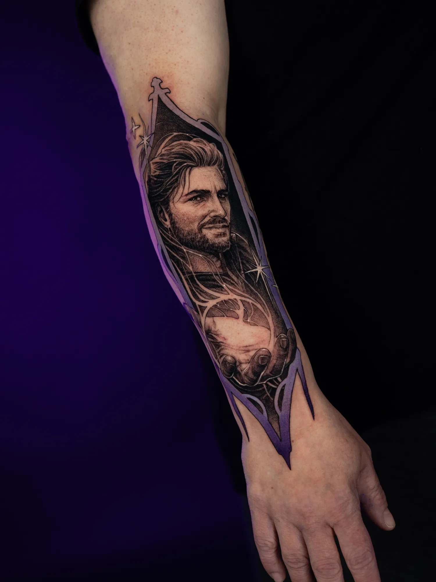 Black and grey portrait tattoo on the outer forearm of Gale from Baldur's Gate III holding a glowing orb, framed by a tall gothic border shaded in purple with small white sparkles.