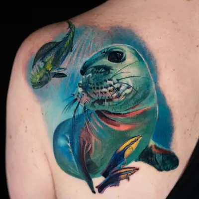 Color realism tattoo on the shoulder blade of a Hawaiian monk seal in blue-green water, with a green and yellow mahi-mahi above it and two small blue and yellow striped fish below.