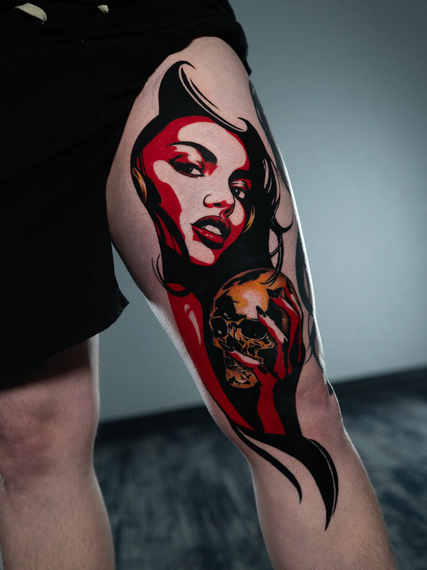 Tattoo of a woman's face with a skull, colored in red and black, on a leg.