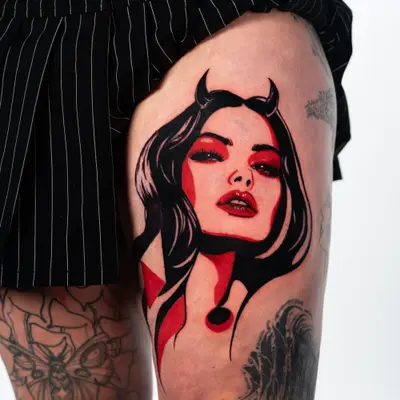 Tattoo of a woman's face with devil horns and red highlights on thigh.