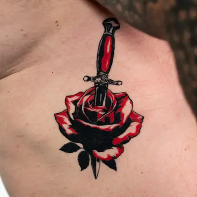 Traditional tattoo of a dagger through a rose in red and black ink.