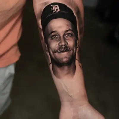 Realistic portrait tattoo on forearm of a man with a cap.