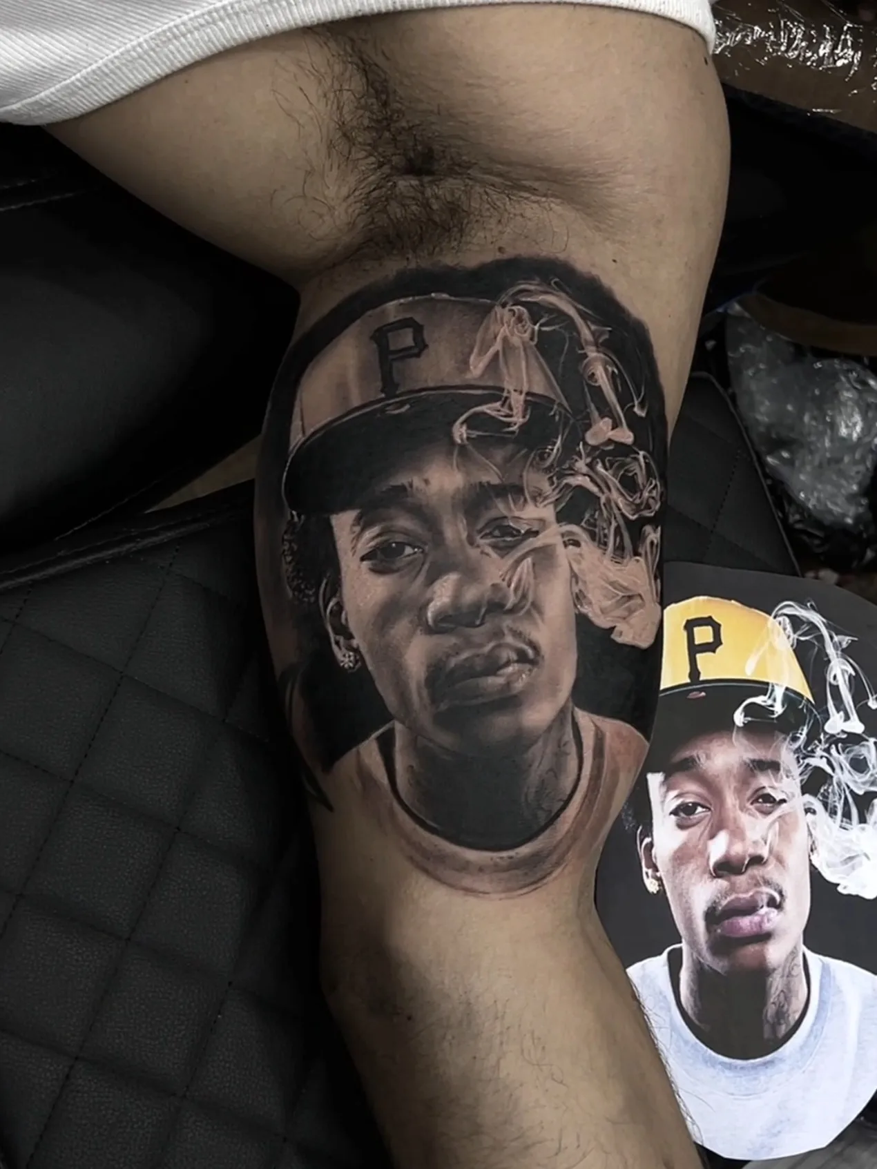 Realistic portrait tattoo on a thigh, showing a man in a cap with smoke around him.