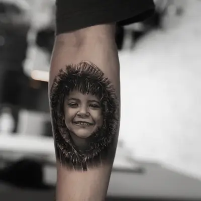 Realistic portrait tattoo of a child in a hoodie on a forearm.