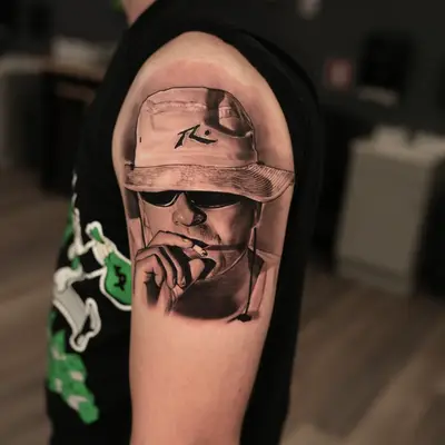 Realistic portrait tattoo of a person wearing a hat and sunglasses on upper arm.