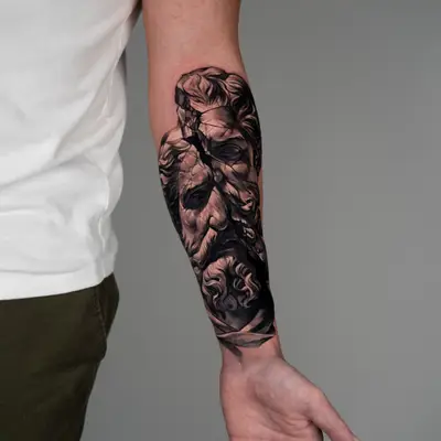 A detailed black and grey tattoo on a forearm featuring a cracked face with elaborate hair.
