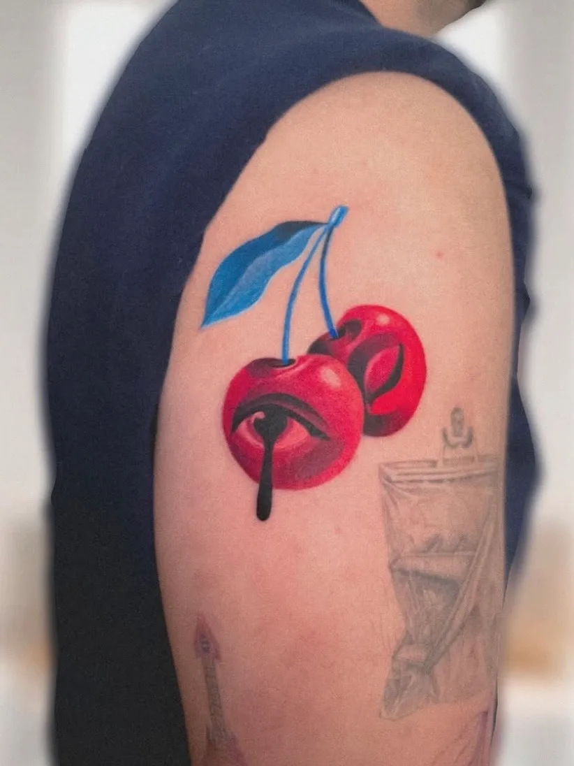 Colorful tattoo of two cherries with an eye design on upper arm.
