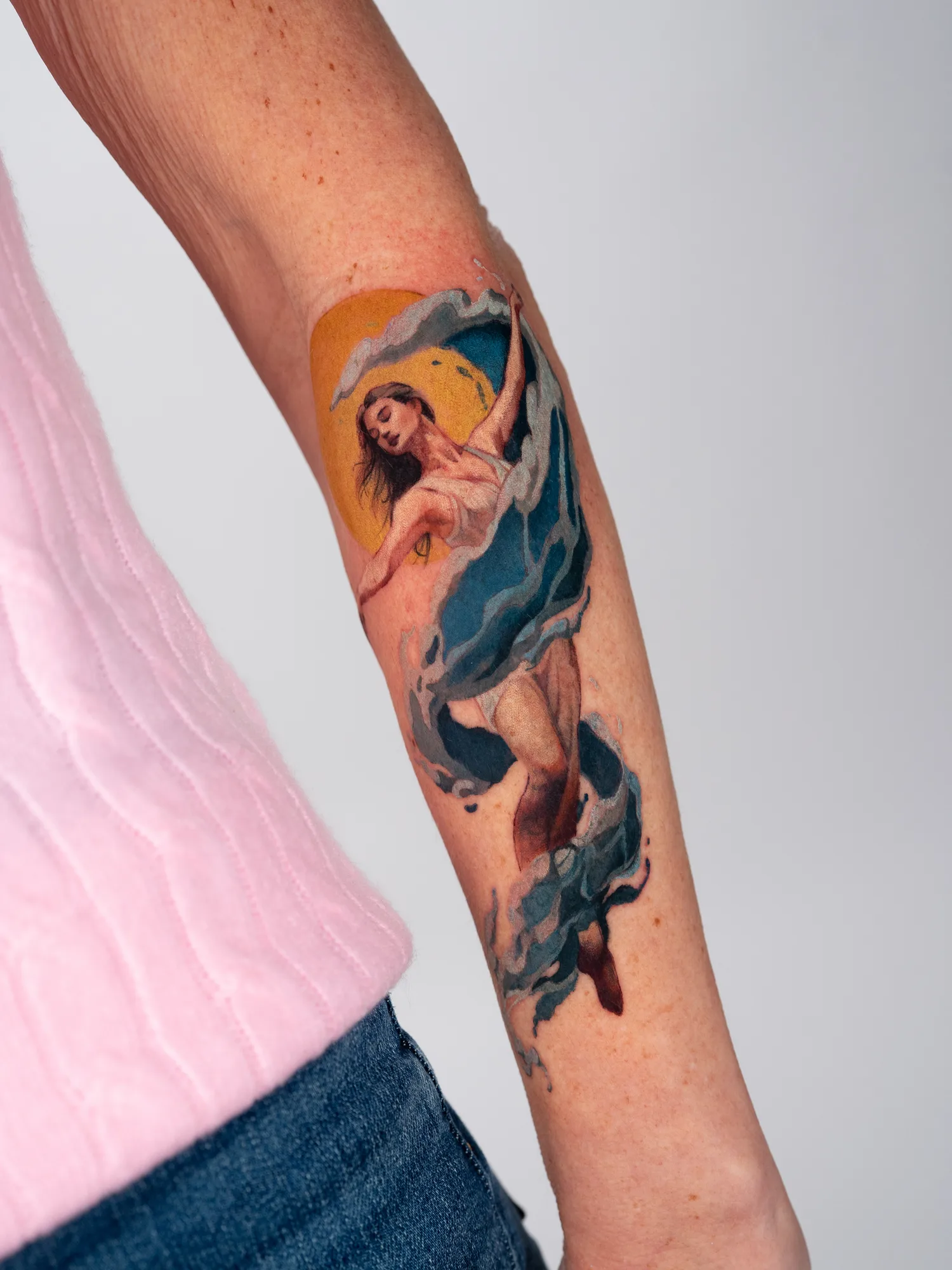 An illustrative realism tattoo on the outer forearm of a woman with flowing wave accents and a sun in the background.