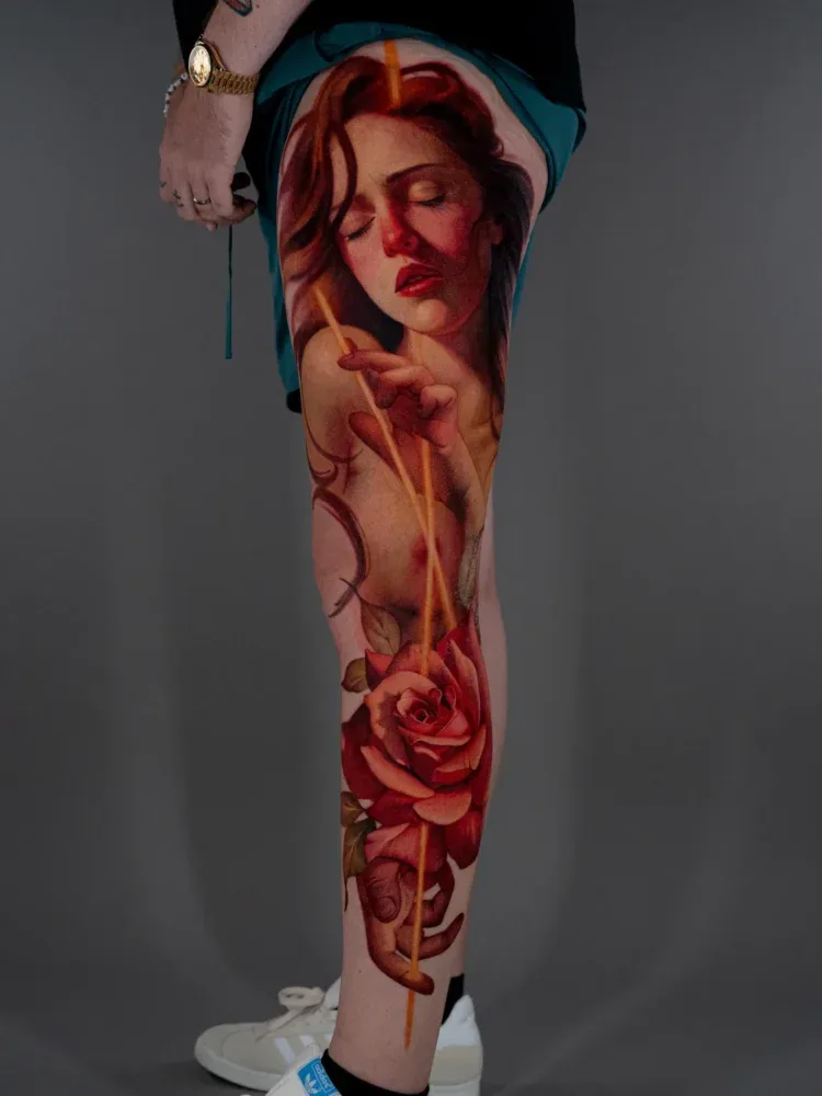 Tattoo of a woman's face with closed eyes and a red rose on the thigh.