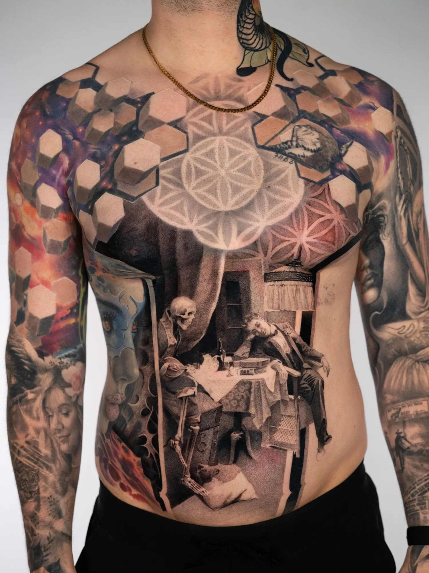 Black and grey stipple tattoo across a stomach showing a skeleton seated at a dinner table with a man in a suit slumped back in his chair, a hanging lamp above and a sacred geometry pattern across the chest.