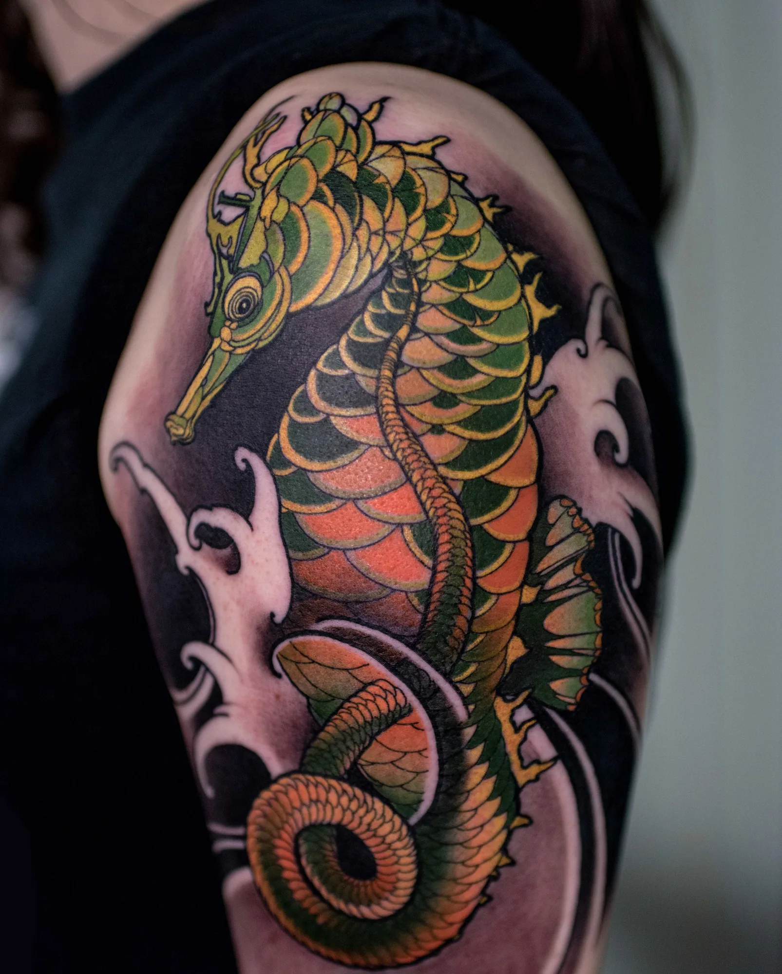 Color neo-traditional seahorse on the upper arm and shoulder, its scales in green, gold and orange with a curled tail, framed by white-capped black waves.