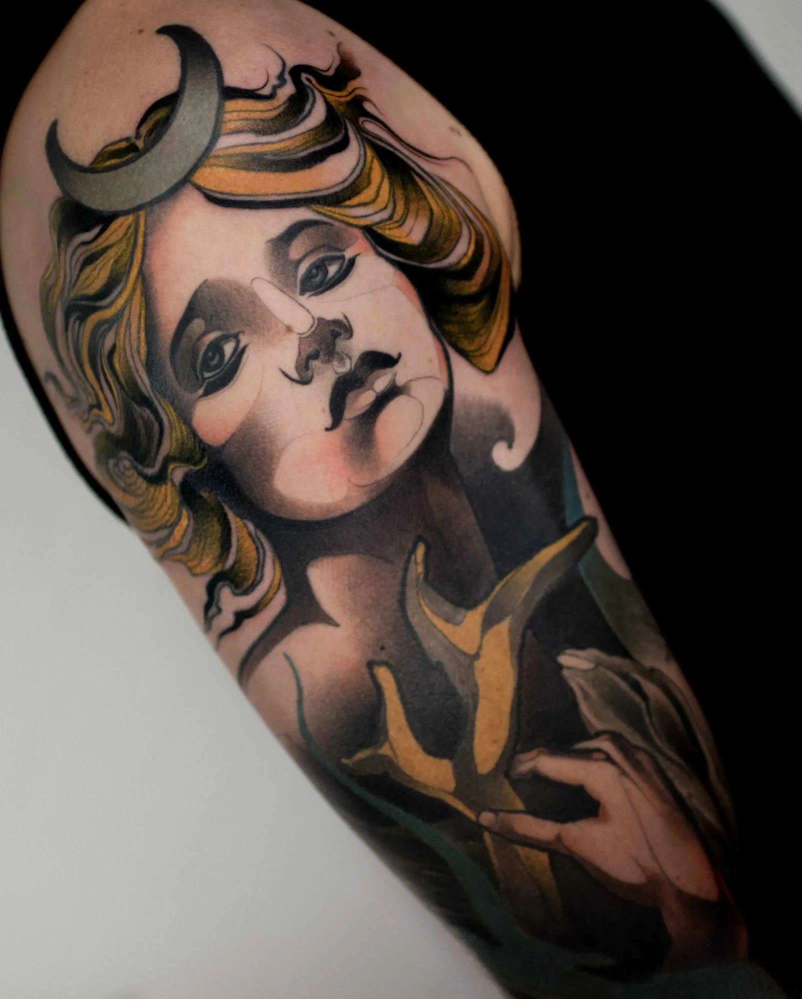 Fusion neo-traditional portrait of a goddess on the upper arm: soft grey face tilted upward, golden wavy hair, a silver crescent moon at the shoulder, and hands holding a golden branching shape below.