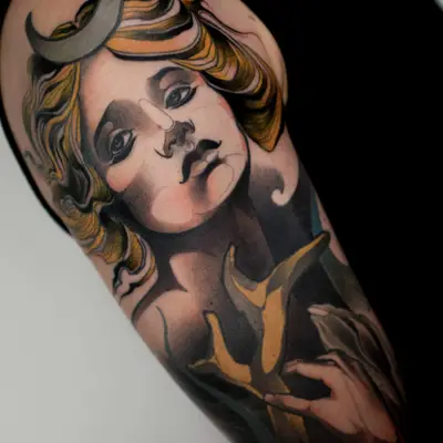 Fusion neo-traditional portrait of a goddess on the upper arm: soft grey face tilted upward, golden wavy hair, a silver crescent moon at the shoulder, and hands holding a golden branching shape below.