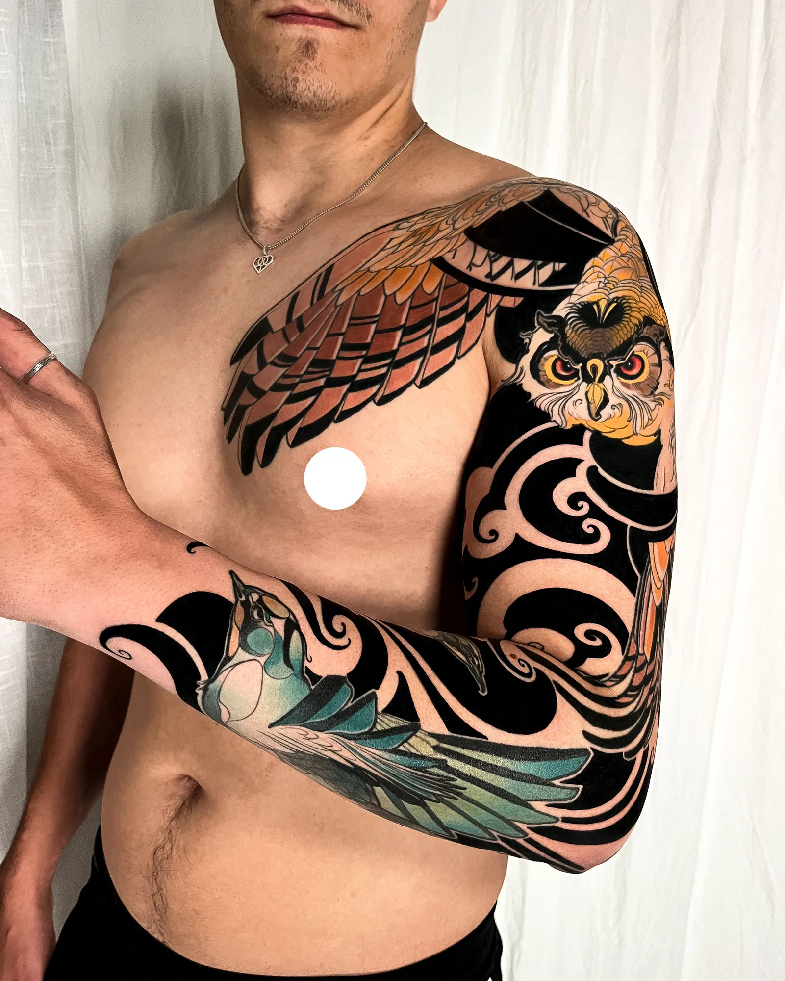 Full color neo-traditional sleeve on a man's left arm: an owl with orange eyes on the shoulder, its brown wing spreading across the chest, and a teal bird on the forearm, all set in bold black ornamental wind and cloud shapes.