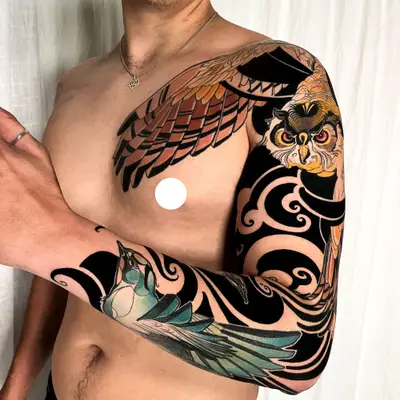 Full color neo-traditional sleeve on a man's left arm: an owl with orange eyes on the shoulder, its brown wing spreading across the chest, and a teal bird on the forearm, all set in bold black ornamental wind and cloud shapes.