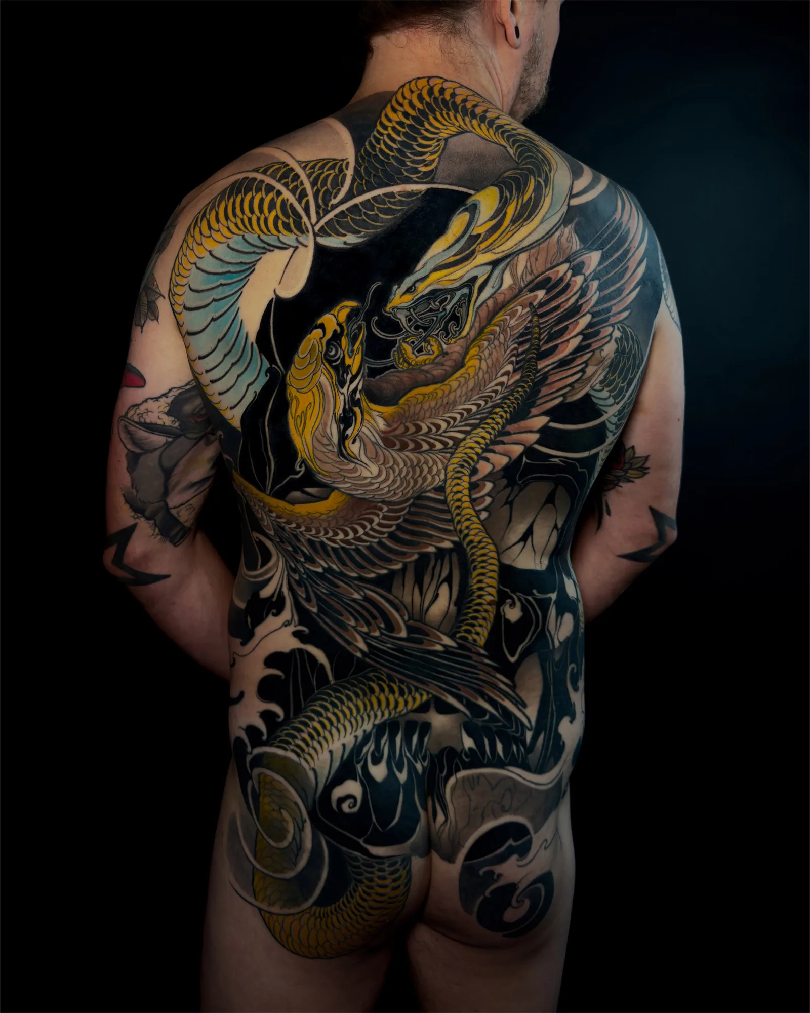 Full back tattoo of a golden snake coiling from the shoulders down to the hips, locked with a winged golden bird across the middle of the back, against a dark background of black wind, flame and wave shapes.