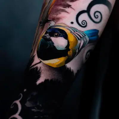 Close-up of a color songbird on a forearm, round body in yellow, navy and white with a bright blue tail, set among black swirls and solid black fill as part of a larger sleeve.