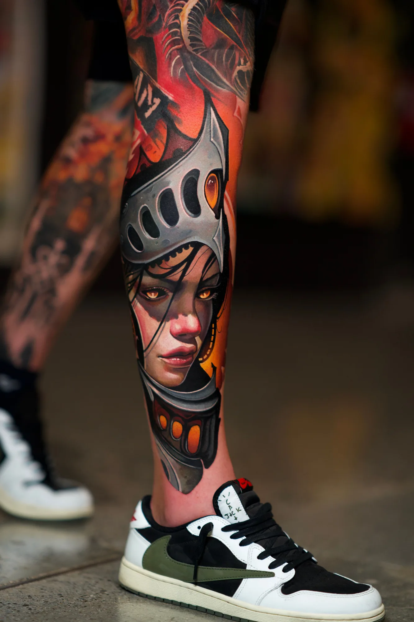 Color realism tattoo on the calf: a woman's face with amber eyes inside an open knight's helmet in steel grey, set against a warm orange and red background that continues up the leg.